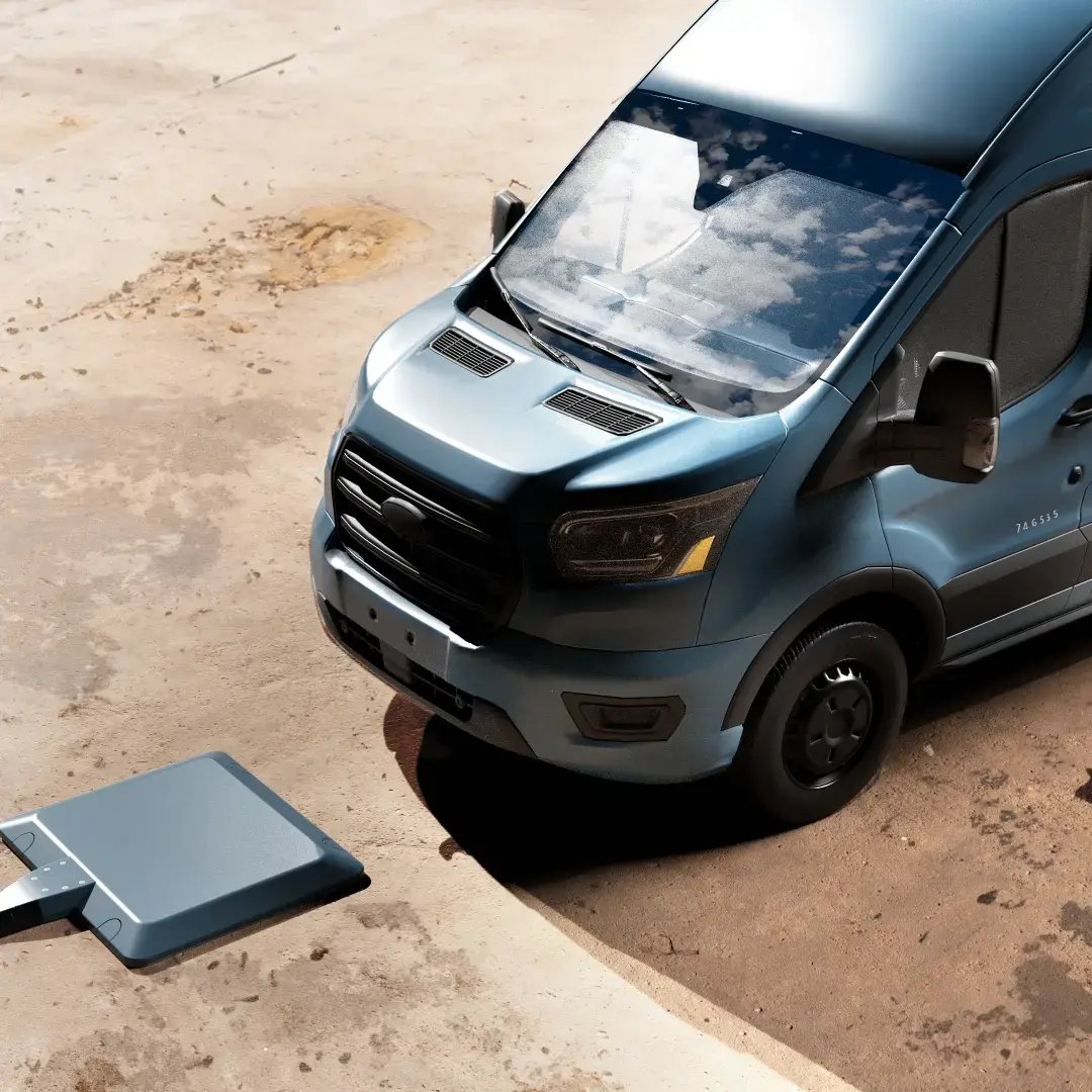 A blue delivery van approaches a wireless charging pad on a rustic parking lot